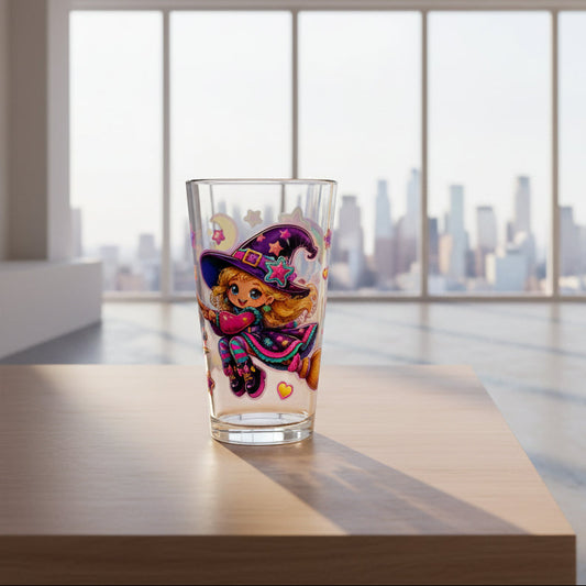A clear pint glass featuring a colorful cartoon illustration of a witch with a purple hat, pink heart, and stars on a white background.