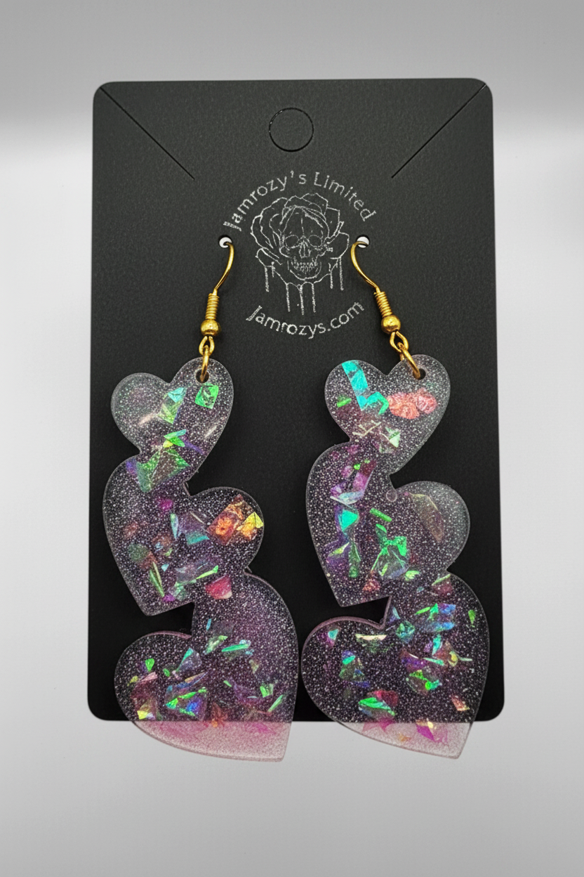 A pair of handmade triple heart-shaped dangle earrings with pink glitter and halo chunks, displayed on a black card with 'Jamrozy's Limited' printed at the top.